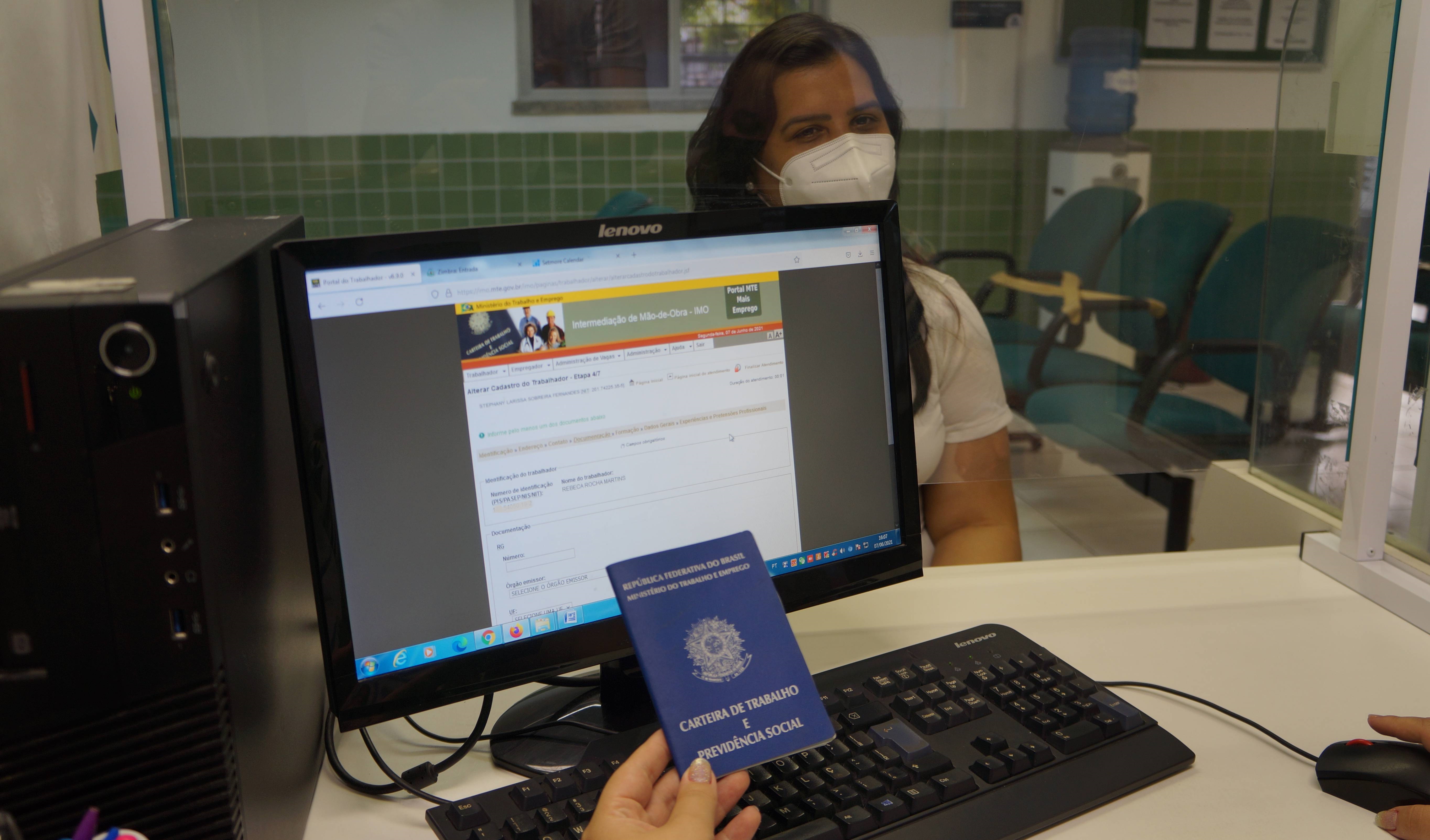 a foto mostra uma mulher sentada num ghichê de atendimento do sine municipal olhando para câmera enquanto a mão de um atendente, que não aparece de corpo inteiro), segura uma carteira de trabalho